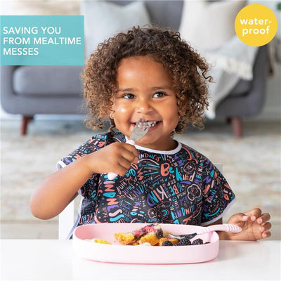 Toddler wearing a colorful Bumkins short sleeve bib, smiling while holding a fork and eating from a pink plate.
