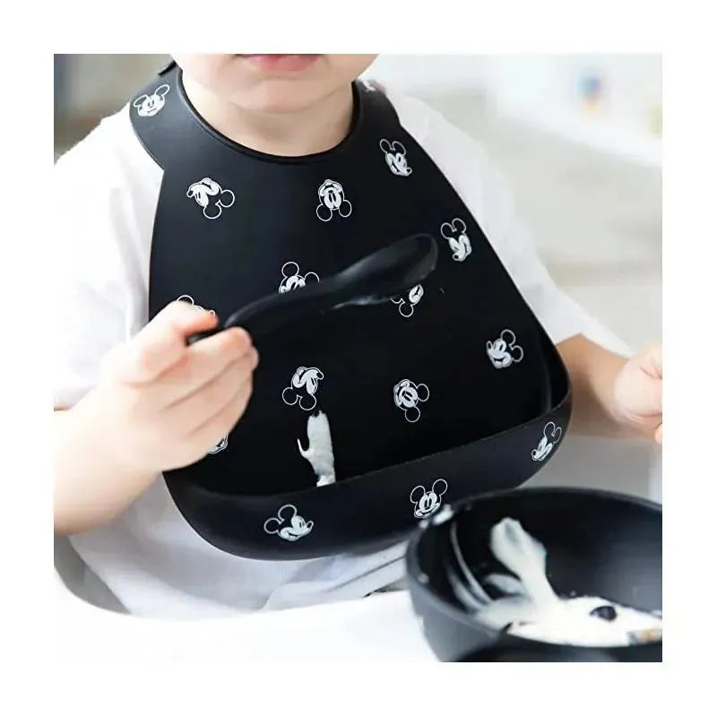 Silicone bib featuring Mickey Mouse designs, worn by a child, with a black spoon and food in a bowl.