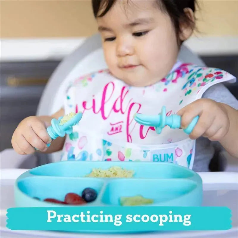 A toddler in a bib uses blue silicone utensils to scoop food from a divided light blue plate, practicing self-feeding.