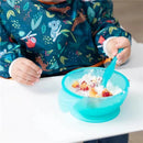 Silicone feeding set in blue with suction cups, contains mixed fruit and yogurt; child using spoon while seated at a table.