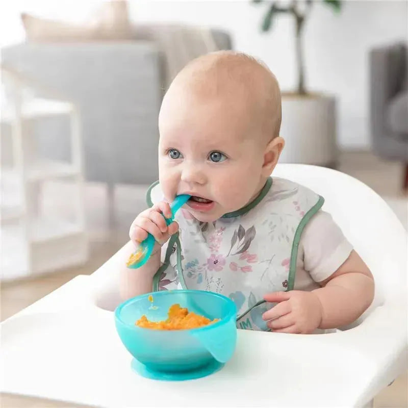 Baby in a high chair using a blue silicone feeding set, including a bowl with food and spoon, wearing a floral bib.
