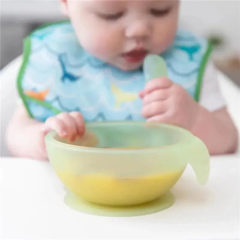 Silicone feeding set in green, featuring a suction bowl and spoon, being used by a baby in a bib.