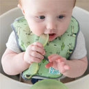 Baby wearing a green Bumkins silicone bib, holding a spoon, with a bowl on a high chair, engaged in feeding.