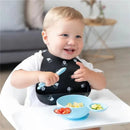 Toddler in a Mickey Mouse bib smiles while using a light blue silicone spoon with a matching feeding set containing food.