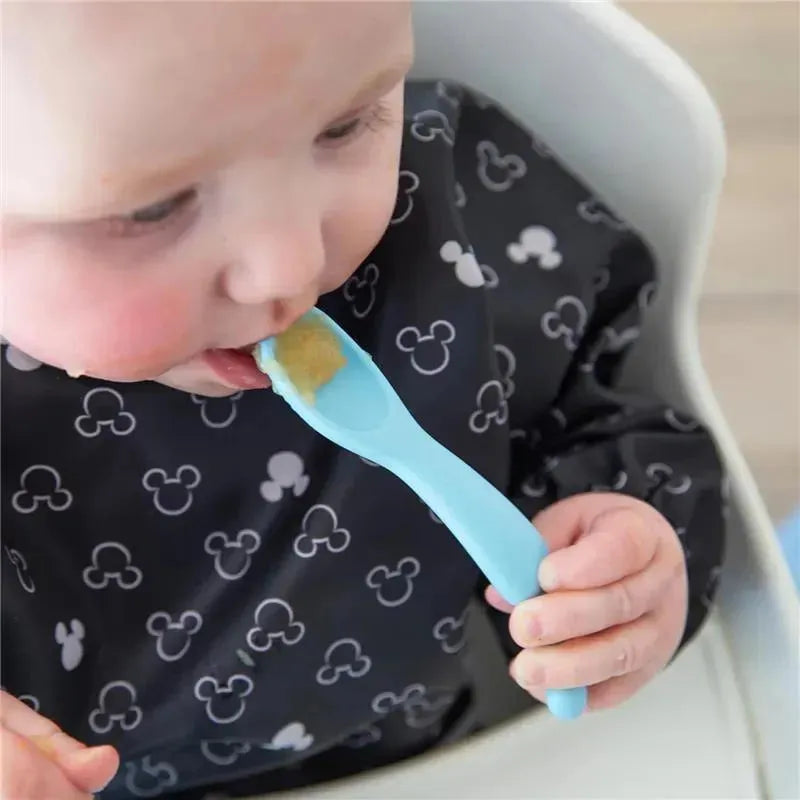 Baby eating with a light blue silicone spoon from a Mickey Mouse-themed feeding set, wearing a waterproof bib.