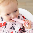 Toddler in a Minnie Mouse bib gnawing on a light pink silicone feeding utensil.