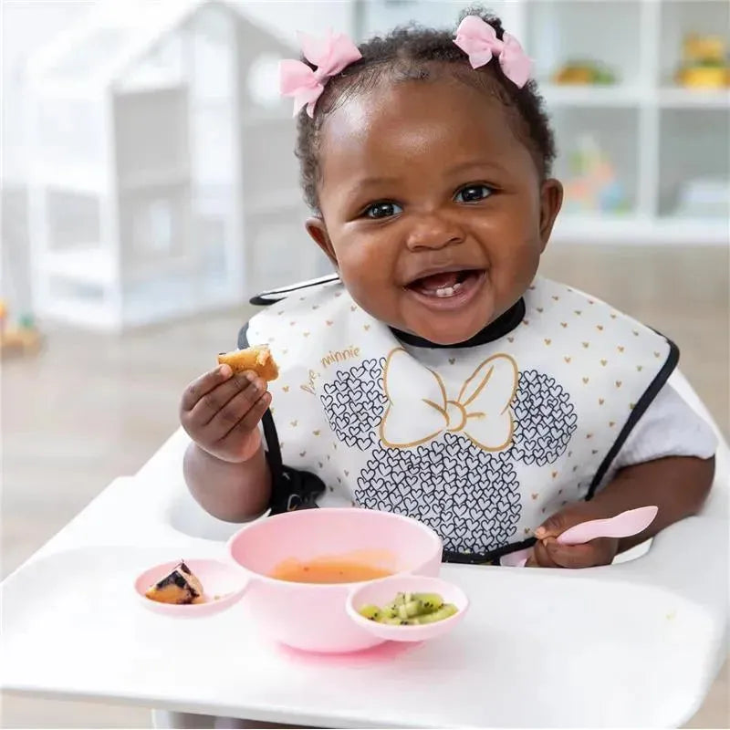 Smiling baby in a Minnie Mouse bib enjoying food, with a pink silicone feeding set and dishes on a high chair tray.