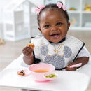 Smiling baby in a Minnie Mouse bib enjoying food, with a pink silicone feeding set and dishes on a high chair tray.