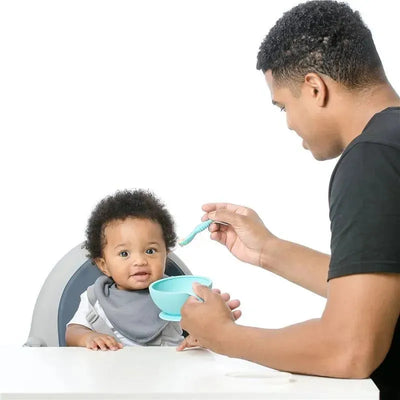 Bumkins silicone first feeding set in blue, featuring a bowl and spoon, with a parent feeding a baby in a high chair.