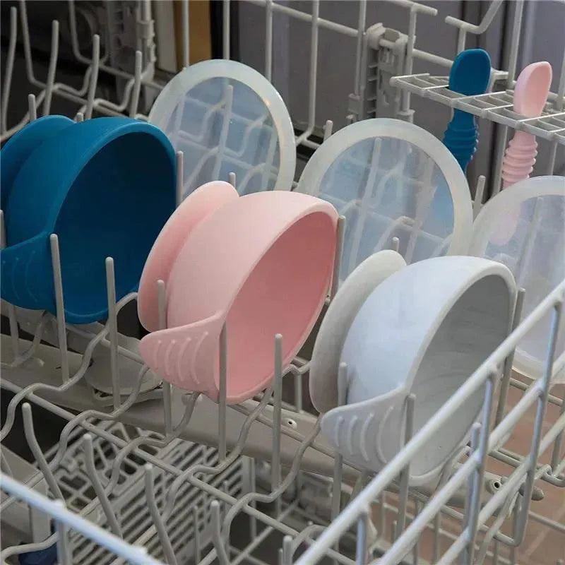 Silicone feeding bowls in pink, blue, and gray with matching spoons, placed in a dishwasher.