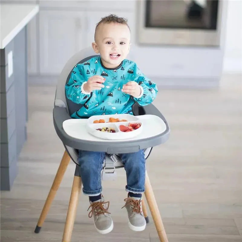 Child wearing a blue bib with a pattern sits in a high chair, smiling, with a silicone grip dish of fruit and snacks on the tray.