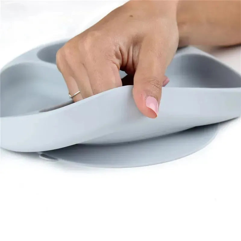 Bumkins gray silicone grip dish with a hand gripping its flexible edge, demonstrating its easy handling feature.