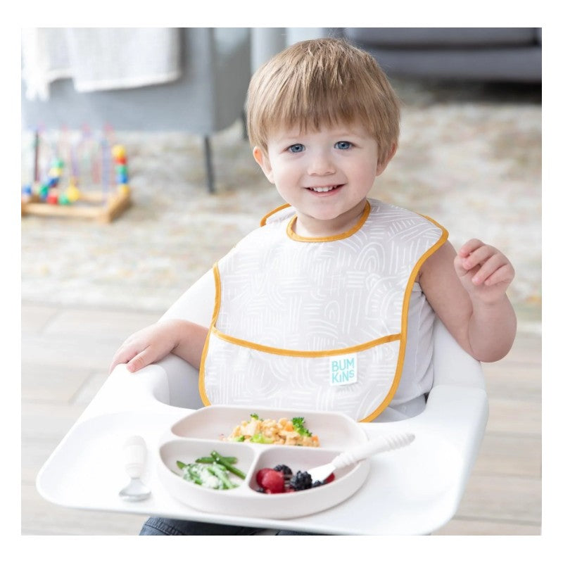 Young child in a highchair wearing a Bumkins bib, smiling while holding a silicone grip dish with divided food sections.