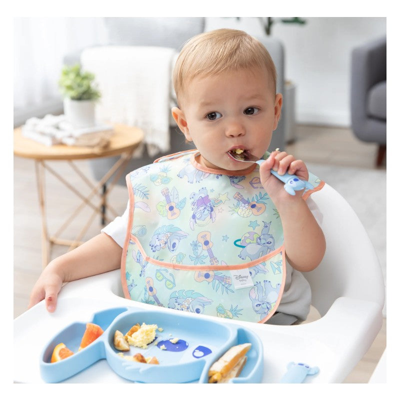 A toddler wearing a Stitch-themed silicone bib eats from a blue grip dish with assorted food items, including fruit.