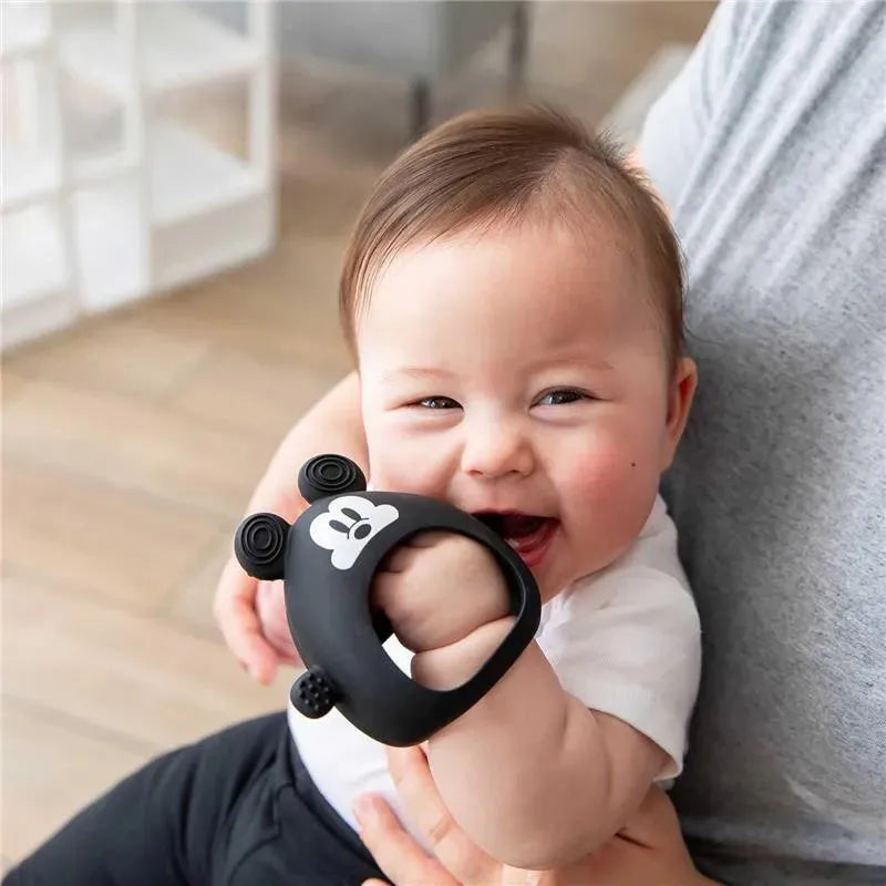 Baby smiling while holding a black silicone mitt shaped like Mickey Mouse from Bumkins.