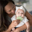 A smiling baby holds a green silicone cactus teether while being gently supported by a caregiver.