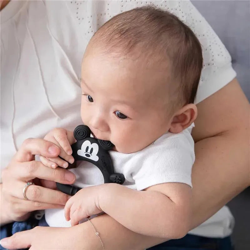 A baby in a white onesie is holding and chewing on a black silicone teether shaped like Mickey Mouse, while being cradled.