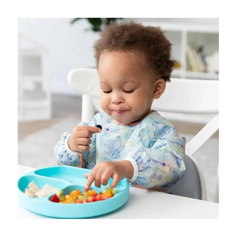 Toddler wearing a Bumkins Aloha sleeved bib, enjoying a colorful meal of fruit and chicken from a blue plate.