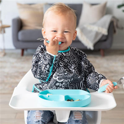 Bumkins Sleeved Bib worn by a happy toddler sitting in a high chair, smiling and holding a spoon with food on a tray.