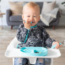 Bumkins Sleeved Bib worn by a happy toddler sitting in a high chair, smiling and holding a spoon with food on a tray.