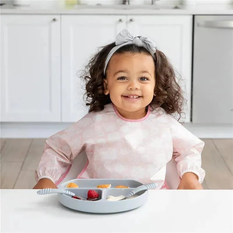 Smiling child wearing a pink Bumkins sleeved bib sits at a table with a gray plate of fruit and yogurt.