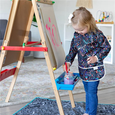A young girl wearing a Bumkins Sleeved Smock decorated with colorful kindness-themed words stands beside an easel, painting.