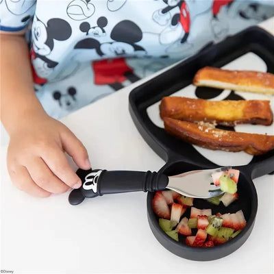 Bumkins Disney Mickey spoon and fork set, with a child using the spoon to eat fruit salad from a bowl on a tray.