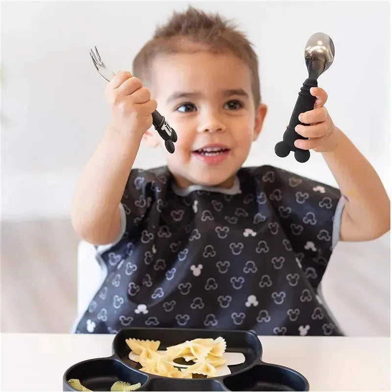 Child in a Mickey Mouse bib happily holds a spoon and fork set while sitting at a table with a divided plate of pasta.