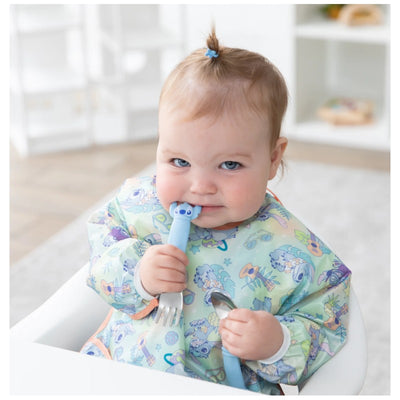 Baby holding a blue Bumkins spoon and fork set while wearing a colorful bib printed with cartoon characters.