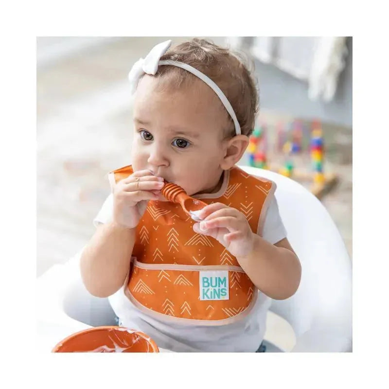 Toddler wearing an orange Bumkins SuperBib with a desert boho pattern, holding a spoon near a bowl of food.
