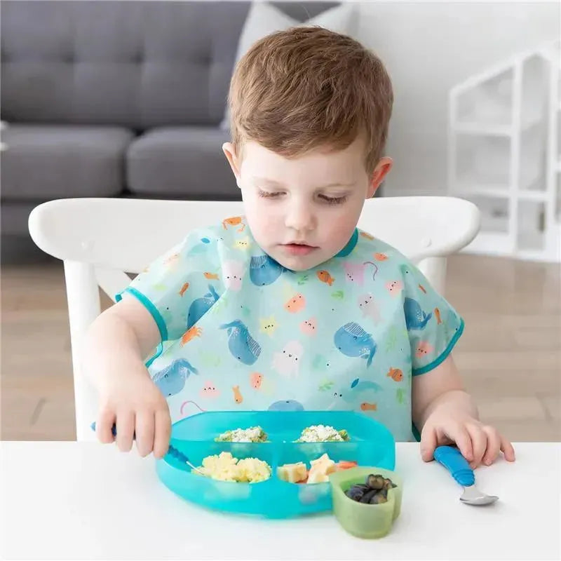 A young child in a colorful bib sits at a table, using a blue silicone grip dish filled with food items.