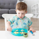 A young child in a colorful bib sits at a table, using a blue silicone grip dish filled with food items.