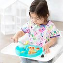 A young child with a colorful floral shirt uses a silicone blue dish to eat scrambled eggs and cherry tomatoes.