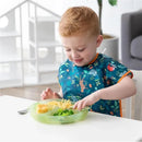 Toddler using a green silicone suction plate with divided sections, filled with pasta, broccoli, and chicken.