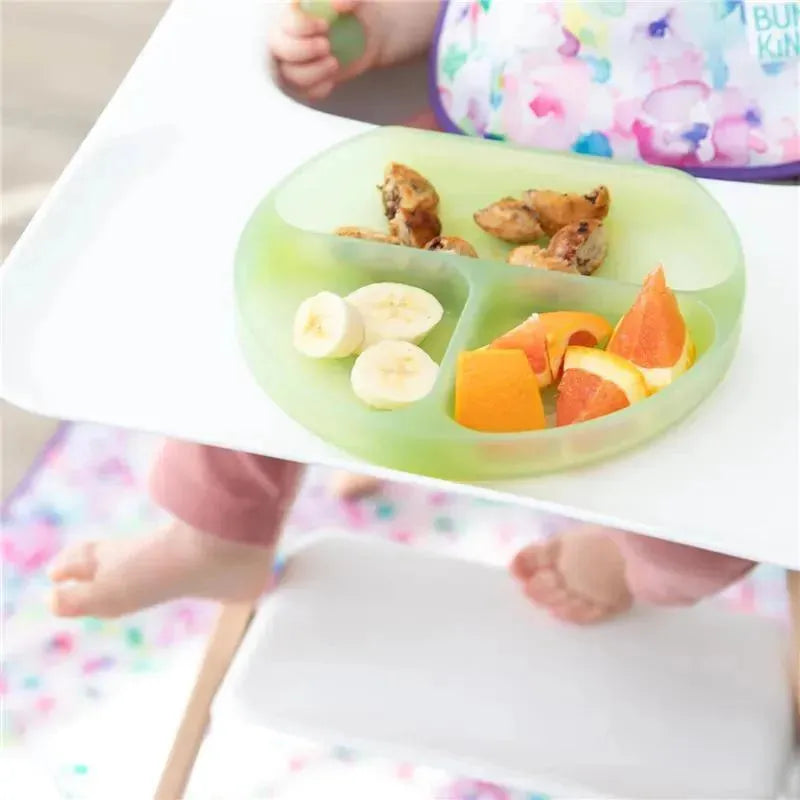 Green silicone suction plate by Bumkins, featuring divided sections with fruit and snacks, held by a toddler.