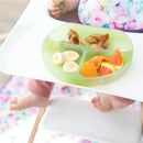 Green silicone suction plate by Bumkins, featuring divided sections with fruit and snacks, held by a toddler.