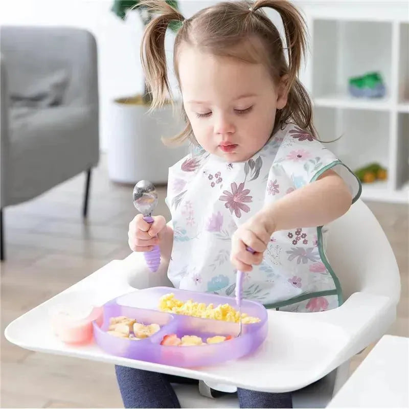 A toddler with pigtails sits in a high chair, using a spoon and fork to eat from a purple silicone grip dish filled with food.