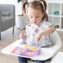 A toddler with pigtails sits in a high chair, using a spoon and fork to eat from a purple silicone grip dish filled with food.