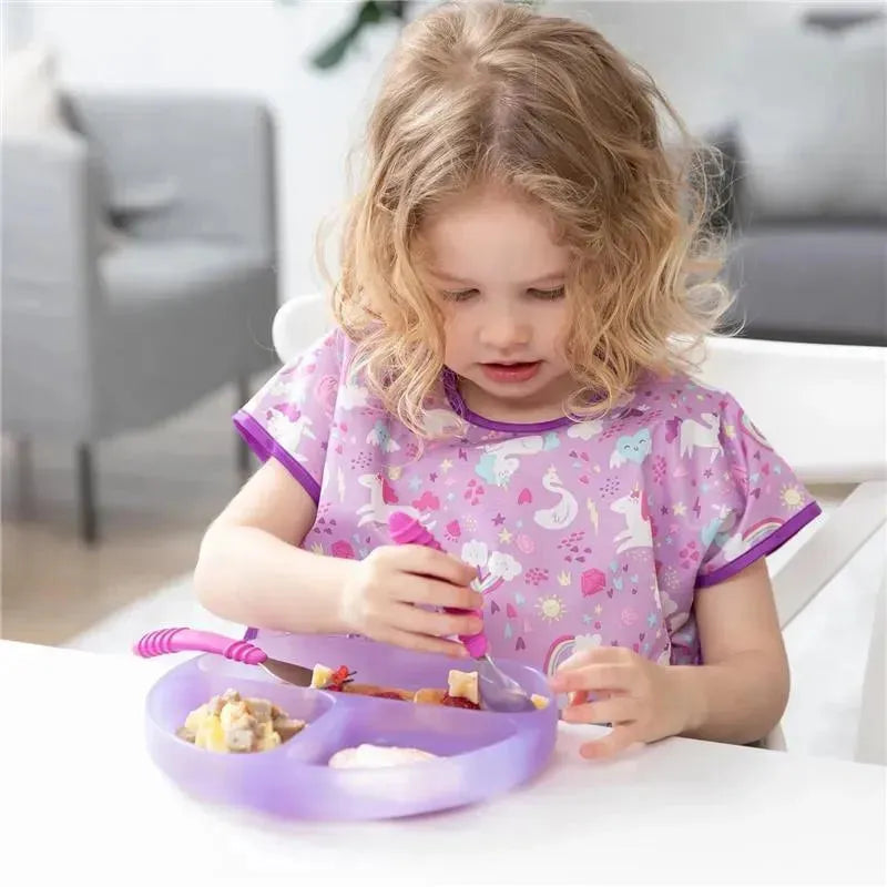 A young child in a purple unicorn-patterned smock uses a fork to eat from a purple silicone grip dish.