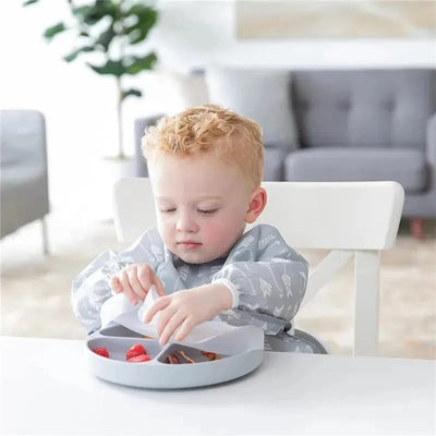 Toddler seated at a table, using a gray Bumkins suction plate with compartments filled with strawberries and snacks.