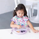 A toddler in a colorful floral bib uses a fork to eat fruit from a lavender-colored suction plate.