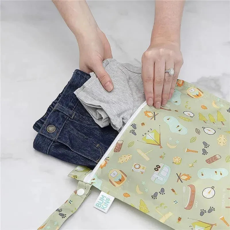 Bumkins waterproof wet bag featuring a camping design, being filled with a gray shirt and denim jeans.