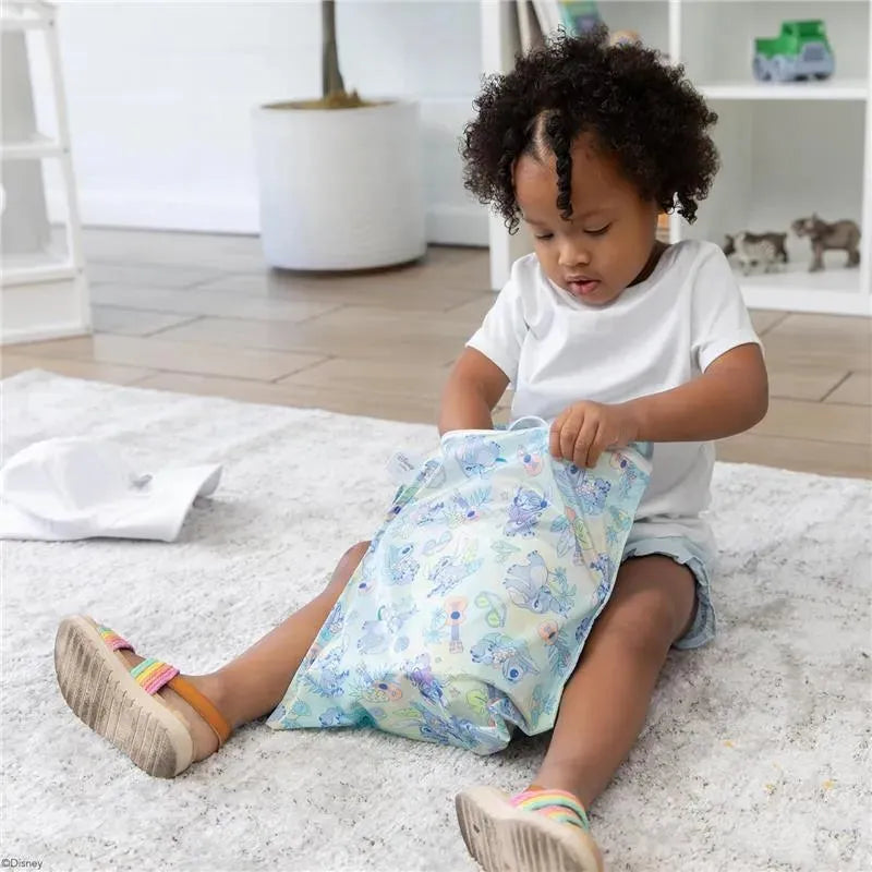 Child in a white shirt sits on a rug, opening a Bumkins Aloha Wet Bag with colorful designs featuring various characters.