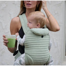 Bumpsuit The Armadillo Baby Carrier in Matcha color, worn by a woman holding a green drink, with a child securely inside.