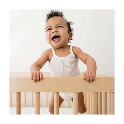 Smiling baby in a white one-piece bodysuit, standing in a wooden crib, beams with joy against a light background.