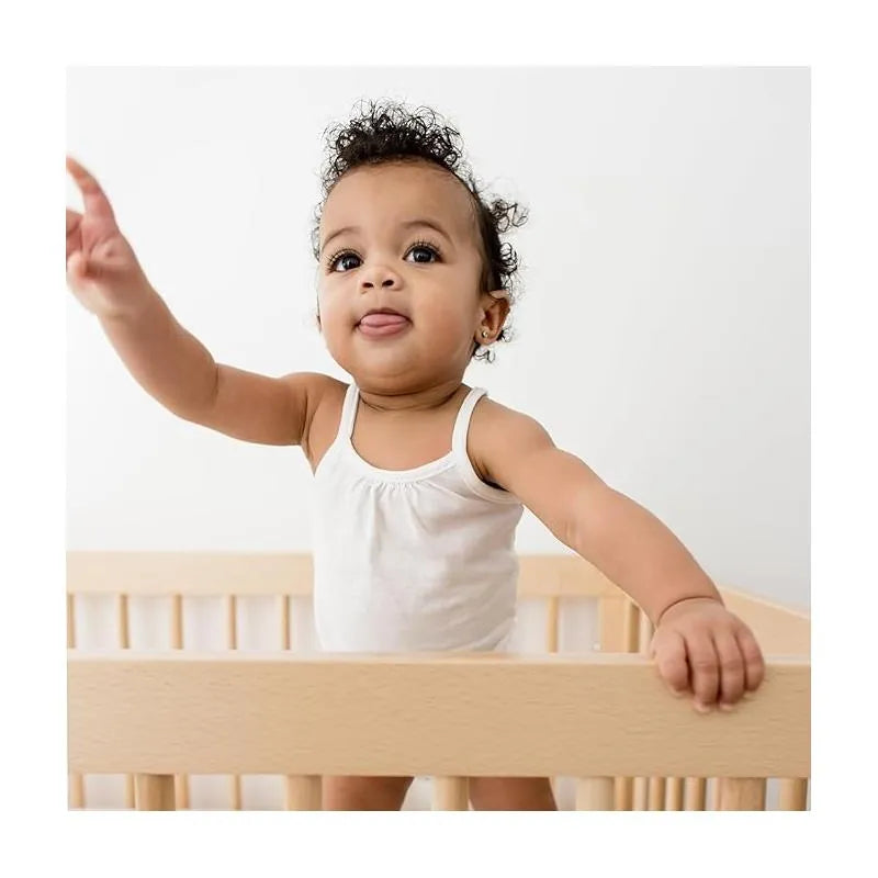 A baby with curly hair, wearing a white tank bodysuit, sits in a wooden crib, raising one arm and sticking out their tongue.