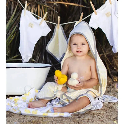 Baby sitting on a blanket with a striped hooded towel, holding bath toys, next to a bowl and hanging baby clothes.