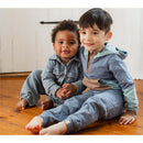 Two young children sit on a wooden floor, wearing matching denim and pastel-colored jumpsuits and smiling at the camera.