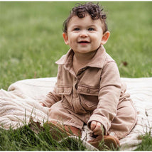 Infant wearing a Burts Bees acid wash jumpsuit in ginger loaf color, smiling while sitting on a blanket in grass.
