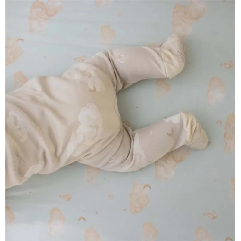 Baby wearing a beige Burts Bees Sleep & Play outfit with cloud patterns, on a light blue sheet with moon graphics.